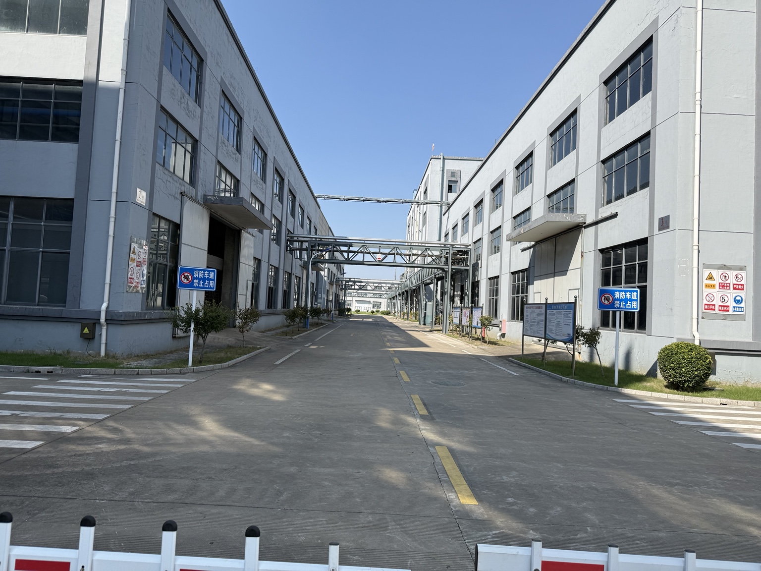 Actual factory exterior with production buildings on both sides of a central roadway and overhead pipe bridges